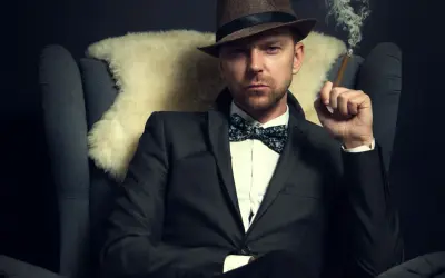 Homme au costume élégant, chapeau et nœud papillon, avec une coupe cheveux rockabilly homme. Il fume un cigare, assis dans un fauteuil.