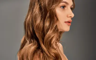 Chevelure ondulée, passant du blond au chatain, d'une femme au profil délicat, sur fond gris.