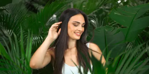 Une jeune femme aux cheveux lisses et sombres, souriant doucement, se tient parmi des feuilles vertes luxuriantes.
