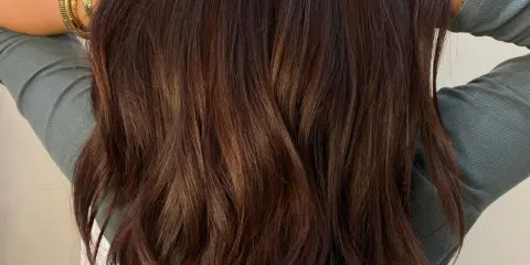 Chevelure brune ondulée, mise en valeur par des reflets subtils. Une femme pose, bras levés, portant des bracelets dorés.