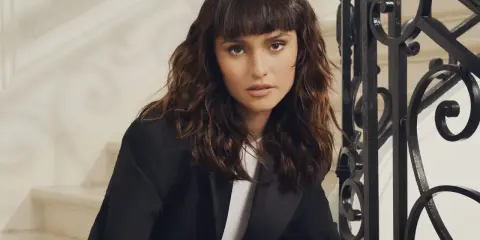 Une femme au regard intense arbore un **wolf cut cheveux bouclés** brun, porté avec un blazer noir.