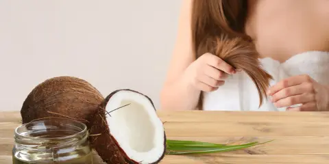 Un pot d'huile de coco et des noix de coco sur une table en bois, avec une femme en arrière-plan qui touche ses cheveux. L'huile de coco est idéale pour les cheveux bouclés.