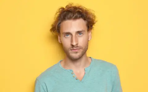 Un jeune homme aux cheveux bouclés et aux yeux bleus regarde la caméra sur fond jaune.