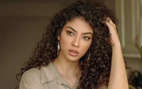 Une jeune femme aux cheveux bruns et bouclés, portant une chemise beige et de grandes boucles d'oreilles. Sa coupe boucles met en valeur son regard intense.