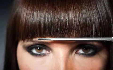 Un modèle de coiffure aux yeux marron et au maquillage prononcé, une frange brune est coupée par des ciseaux.
