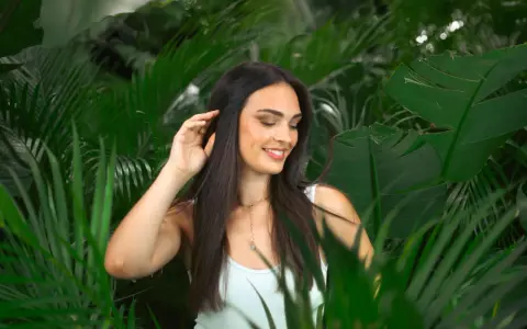 Une jeune femme aux cheveux lisses et sombres, souriant doucement, se tient parmi des feuilles vertes luxuriantes.