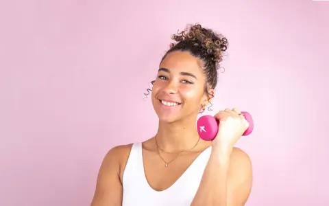 Une jeune femme souriante avec une coiffure sport cheveux bouclés tient une haltère rose.