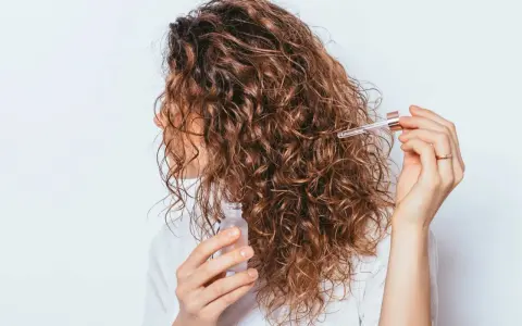 Une femme applique une huile capillaire sur ses cheveux frisottis sur le dessus. Ses boucles brunes brillent sous la lumière.