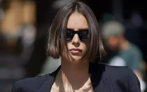 Une femme au regard intense arbore une coupe carré droit dégradé, accentuée par des lunettes de soleil noires et un blazer chic.