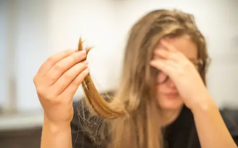 Une main tient une mèche de cheveux, visiblement abîmés, comme si c'était du cheveux mort. La personne semble désespérée.