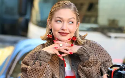 Gigi Hadid, avec ses boucles d'oreilles en forme de dé rouge, pose avec un sourire malicieux. Son carré plaqué en arrière et son manteau texturé complètent son look.