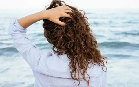 Main tenant ses cheveux bouclés, une femme en chemise rayée profite de la brise marine. Elle n'a pas peur de brosser ses cheveux bouclés tous les jours.