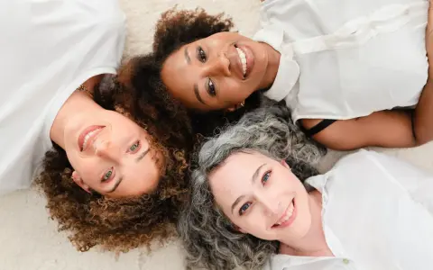 Trois femmes souriantes, aux cheveux bouclés, frisés et gris, partagent un moment complice. Un scruncher cheveux pourrait parfaire leur look.