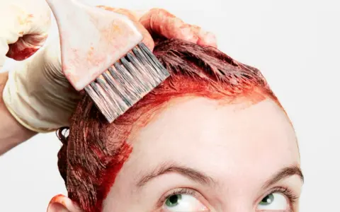 Application de teinture rouge sur les cheveux, étape clé de la routine cheveux colorés. Une main gantée utilise un pinceau pour une application précise.