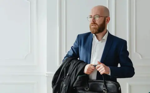 Un homme crane rasé, barbe rousse, lunettes, en costume bleu, porte une veste noire et une mallette.