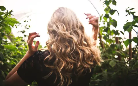 Chevelure blonde ondulée, baignée de lumière, entourée de verdure. Une scène évoquant la douceur d'une **décoloration végétale** naturelle.