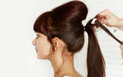 Une main ajuste une mèche pour créer un chignon sophistiqué. La coiffure volumineuse est en cours de réalisation.