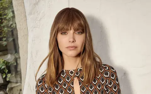 Une femme au carré frange rideau brune, aux yeux verts perçants, porte une robe à motifs géométriques.