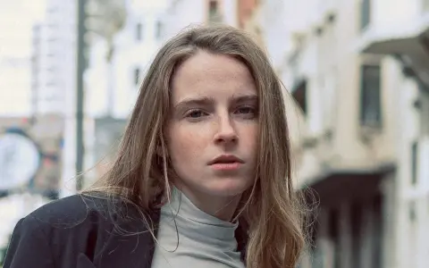 Jeune femme au regard intense, ses cheveux châtain clair cendré ou glacé encadrent son visage parsemé de taches de rousseur. Elle porte un col roulé gris et une veste sombre.