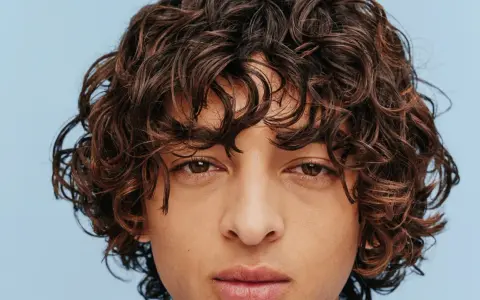 Un jeune homme au regard intense arbore une magnifique coupe cheveux bouclés homme, des boucles brunes et rebelles encadrant son visage.