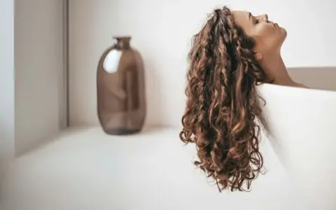 Une femme aux cheveux bouclés se détend dans une baignoire. Elle se demande combien de shampoing par lavage est idéal pour ses cheveux.