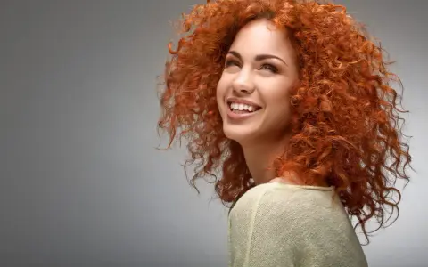 Une jeune femme au dégradé cheveux bouclés mi long, rousse, sourit joyeusement.