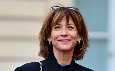 Une femme au sourire radieux arbore une coupe courte femme tendance, des lunettes sur la tête et des créoles argentées.