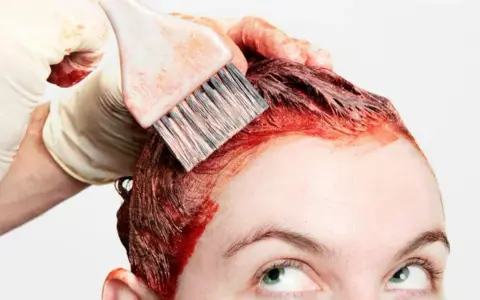 Application d'une teinture rouge pour un nouvel effet couleur cheveux. Une main gantée utilise un pinceau pour colorer les racines.