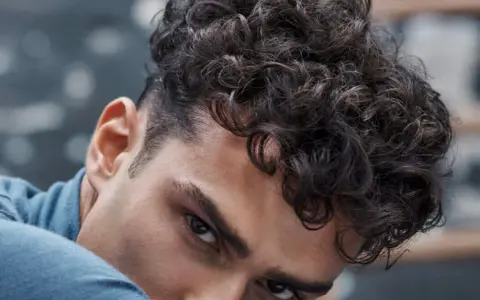 Gros plan sur un homme avec une coupe homme mi long bouclé, ses boucles sombres encadrant son visage.