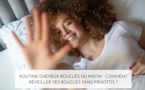 Une femme souriante aux cheveux 3c bouclés se réveille dans son lit. Sa main est devant l'objectif.