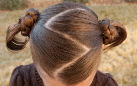 Cheveux bruns tressés en deux couettes rondes, séparés par une raie zig zag.