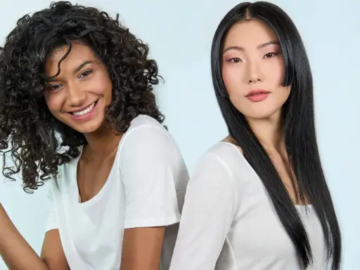 Deux femmes souriantes, l'une aux cheveux bouclés, l'autre aux cheveux lisses. La première joue avec ses mèches, évitant les cheveux fourchus jusqu'à la racine.