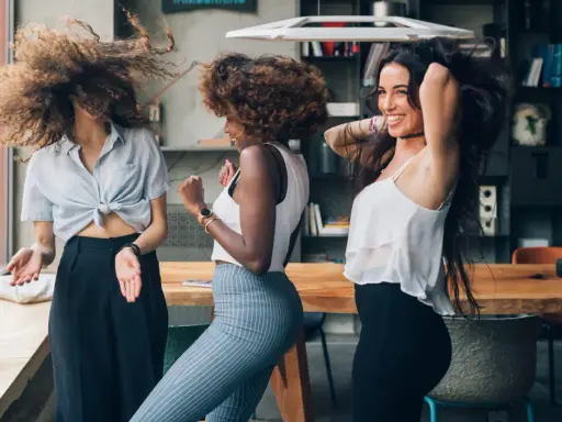 Trois femmes aux cheveux bouclés dansent joyeusement. Leurs chevelures volent, un exemple de comment avoir de belles boucles sans frisottis.