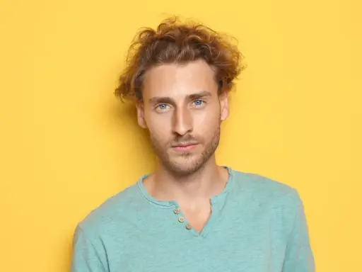 Un jeune homme aux cheveux bouclés et aux yeux bleus regarde la caméra sur fond jaune.