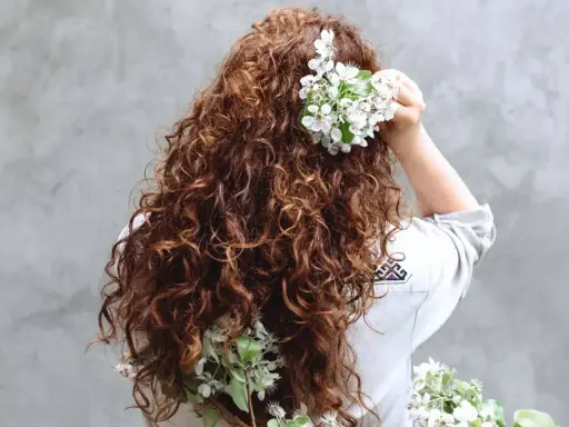 Chevelure brune et bouclée ornée de fleurs blanches, évoquant un décollement de racine naturel et printanier.