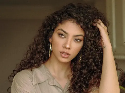 Une jeune femme aux cheveux bruns et bouclés, portant une chemise beige et de grandes boucles d'oreilles. Sa coupe boucles met en valeur son regard intense.