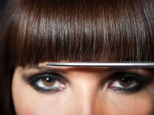 Un modèle de coiffure aux yeux marron et au maquillage prononcé, une frange brune est coupée par des ciseaux.