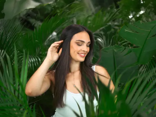 Une jeune femme aux cheveux lisses et sombres, souriant doucement, se tient parmi des feuilles vertes luxuriantes.