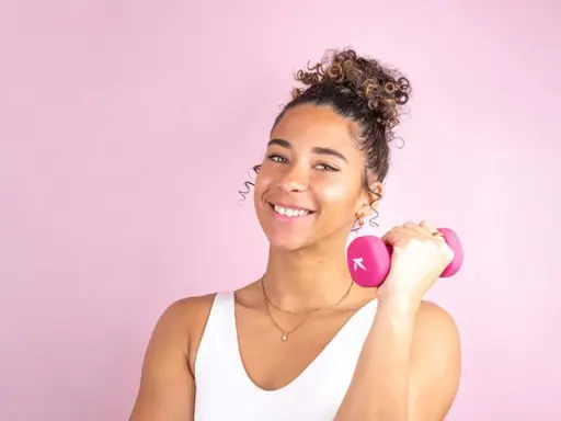 Une jeune femme souriante avec une coiffure sport cheveux bouclés tient une haltère rose.