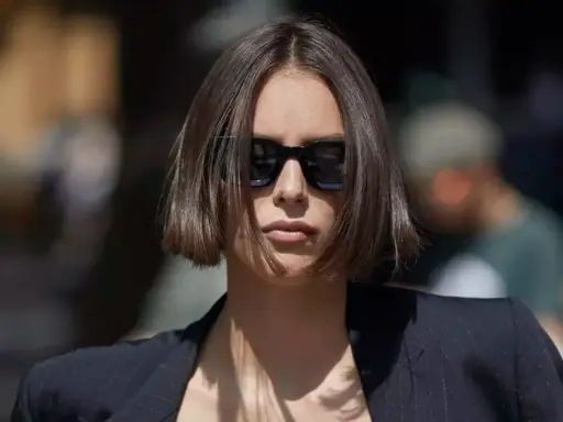 Une femme au regard intense arbore une coupe carré droit dégradé, accentuée par des lunettes de soleil noires et un blazer chic.