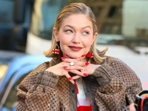 Gigi Hadid, avec ses boucles d'oreilles en forme de dé rouge, pose avec un sourire malicieux. Son carré plaqué en arrière et son manteau texturé complètent son look.