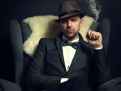 Homme au costume élégant, chapeau et nœud papillon, avec une coupe cheveux rockabilly homme. Il fume un cigare, assis dans un fauteuil.