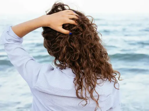 Main tenant ses cheveux bouclés, une femme en chemise rayée profite de la brise marine. Elle n'a pas peur de brosser ses cheveux bouclés tous les jours.
