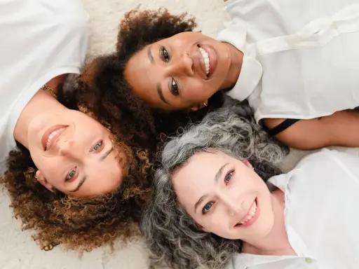 Trois femmes souriantes, aux cheveux bouclés, frisés et gris, partagent un moment complice. Un scruncher cheveux pourrait parfaire leur look.