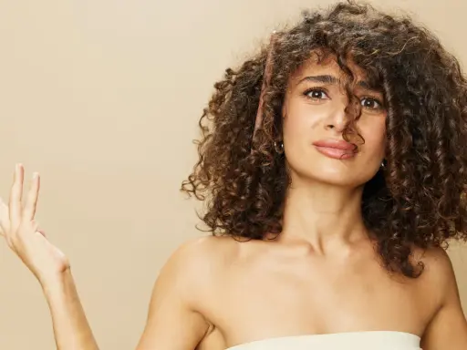 Une femme aux cheveux longs et bouclés, une brosse à cheveux coincée dedans, lève une main ouverte.