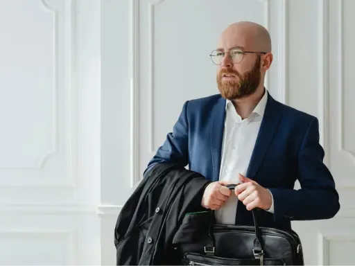 Un homme crane rasé, barbe rousse, lunettes, en costume bleu, porte une veste noire et une mallette.