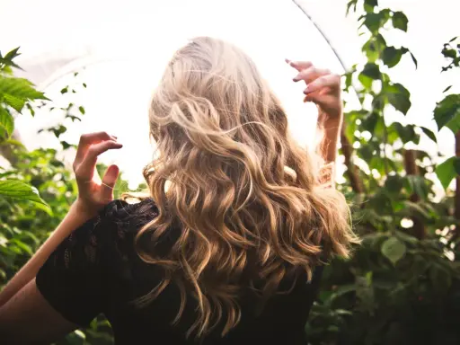 Chevelure blonde ondulée, baignée de lumière, entourée de verdure. Une scène évoquant la douceur d'une **décoloration végétale** naturelle.