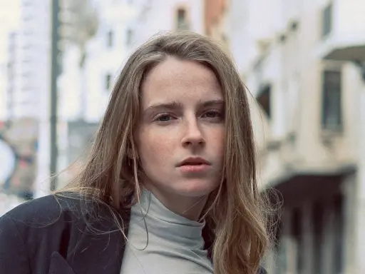 Jeune femme au regard intense, ses cheveux châtain clair cendré ou glacé encadrent son visage parsemé de taches de rousseur. Elle porte un col roulé gris et une veste sombre.