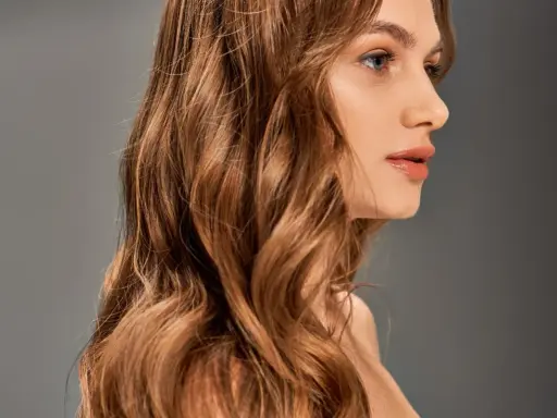 Chevelure ondulée, passant du blond au chatain, d'une femme au profil délicat, sur fond gris.