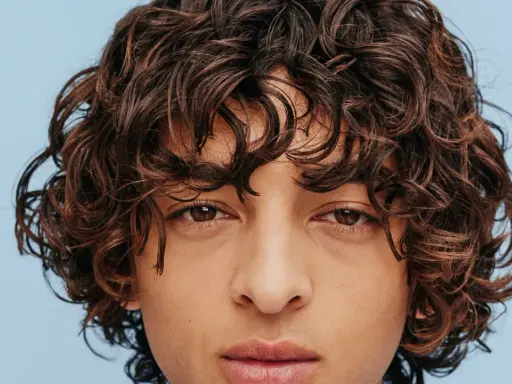 Un jeune homme au regard intense arbore une magnifique coupe cheveux bouclés homme, des boucles brunes et rebelles encadrant son visage.